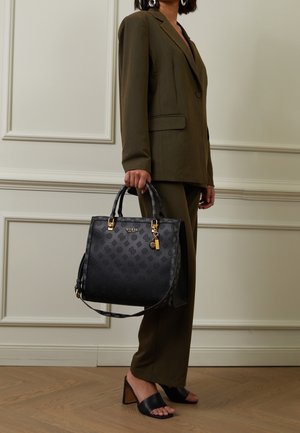 Bolso de mano de cuero negro con patrón de logo en relieve, herrajes dorados y una correa desmontable. Llevado con un traje de color verde oliva.