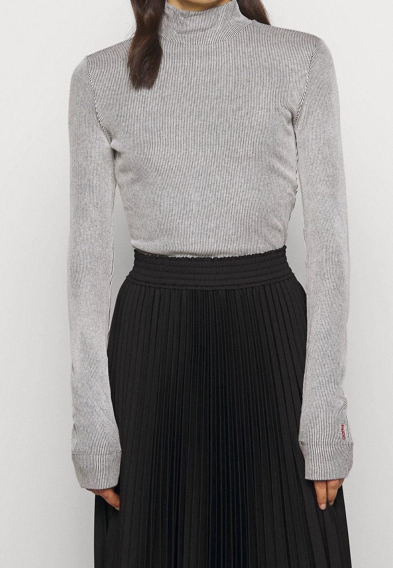 Femme portant un pull à col roulé côtelé gris à manches longues et une jupe noire plissée taille haute, debout devant un fond uni.