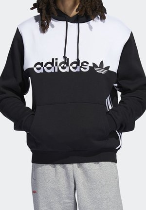Osoba w czarno-białej bluzie Adidas z logo na klatce piersiowej i jasnoszarych spodniach dresowych, ręce włożone do kieszeni bluzy.