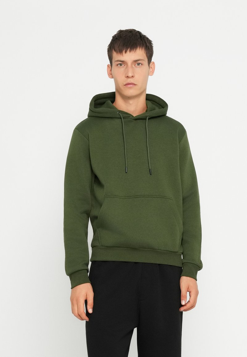 Felpa verde in cotone con tasca a kangaroo, cappuccio regolabile con coulisse e polsini a costine. Indossata con pantaloni neri. Design minimalista.