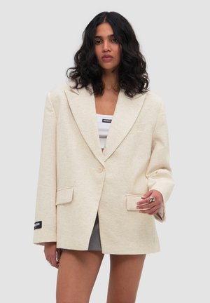 Vrouw met krullend zwart haar, die een oversized beige blazer, een wit cropped topje, een grijze rok en ringen draagt op een lichtgrijze achtergrond.
