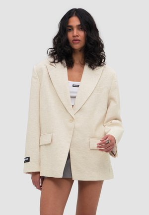 Femme aux cheveux noirs bouclés portant un blazer beige oversize, un crop top blanc, une jupe grise et des bagues sur un fond gris clair.