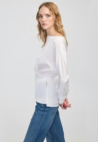 Blonde vrouw met losse golven in een witte blouse met lange mouwen en een blauwe jeans, zijwaarts staand met haar handen achter haar rug gekruist.