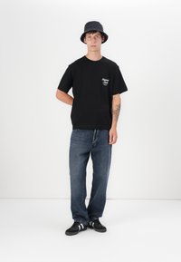 Black t-shirt with a pocket logo, paired with blue denim jeans. Model wears a navy bucket hat and black shoes with white stripes.