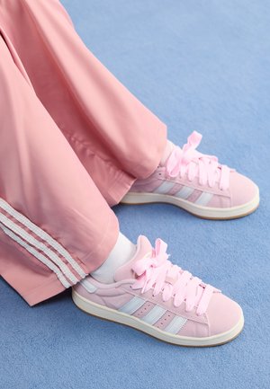 Osoba nosząca różowe buty sportowe Adidas z białymi paskami i różowymi sznurówkami, połączone z różowymi spodniami na jasnoniebieskim dywanie.
