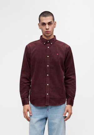 Jeune homme aux cheveux courts portant une chemise en velours côtelé rouge foncé boutonnée et un jean bleu clair, debout devant un fond blanc uni.