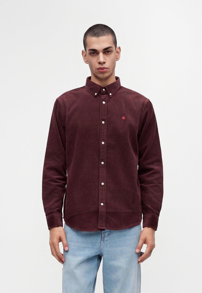 Jeune homme aux cheveux courts portant une chemise en velours côtelé rouge foncé boutonnée et un jean bleu clair, debout devant un fond blanc uni.