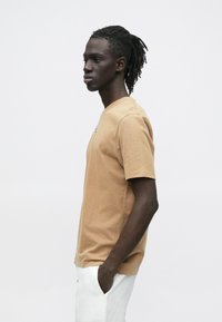 Brun bomuld T-shirt med rund hals, korte ærmer og en afslappet pasform. Modellen står i profil med hænderne i lommerne mod en hvid baggrund.