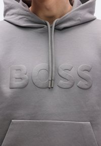 Hall hallipluus pehme tekstuuriga, esiküljel reljeefsed tähed "BOSS" ning paelaga kinnitusega. Sisaldab esitaskut.