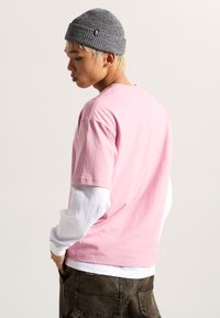 Camiseta rosa de manga corta superpuesta sobre una camiseta blanca de manga larga, combinada con pantalones cargo oscuros y texturizados, y un gorro de punto gris.