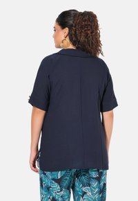 Marineblaue Kurzarmbluse aus weichem Material mit Kragen. Verziert mit dekorativem goldenen Zubehör an den Ärmelbündchen. Getragen mit gemusterten Blumenhosen.