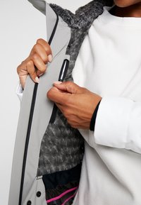 Grå jacka med en krage i pälsfoder, med en dragkedjefick inuti i ett mönstrat tyg. Hand som greppar dragkedjan.