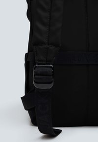 Zaino nero in nylon morbido, con tracolla regolabile e imbottita, e un logo ben visibile su una banda. Include una chiusura sicura con fibbia.