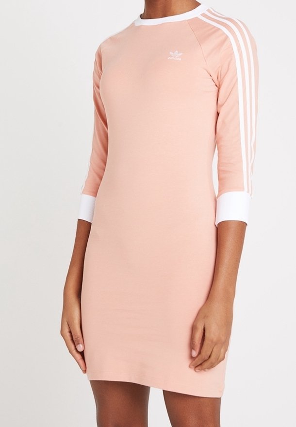 Kvinna som bär en ljusrosa Adidas-klänning med trekvartsärmar som har vita ränder och vita avslut.
