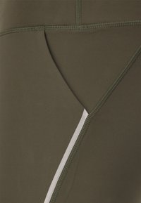 Lähikuva oliivinvihreästä kankaasta, jossa on yksityiskohtainen ompelu ja diagonaalinen heijastava hopea raita sileällä materiaalilla.