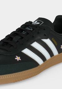 Μαύρο αθλητικό παπούτσι Adidas Samba με λευκές ρίγες, λαστιχένια σόλα, μικρή ροζ κεντημένη λουλουδάκη και χρυσή επιγραφή "SAMBA" στο πλάι.