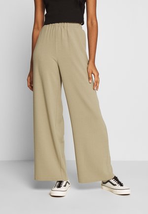 Osoba ubrana w beżowe spodnie z szerokimi nogawkami i elastycznym pasem, czarny top bez rękawów oraz białe trampki z czarnymi sznurowadłami, stojąca na szarej podłodze.