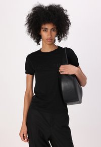 T-shirt nera a maniche corte con una leggera lucentezza e logo in rilievo sul petto, abbinata a una elegante borsa in pelle nera.