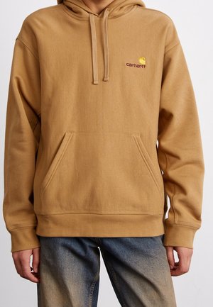 Beige Carhartt hoodie med frontlomme og hættesnore, båret med falmede blå jeans, der viser overkrop og hænder.