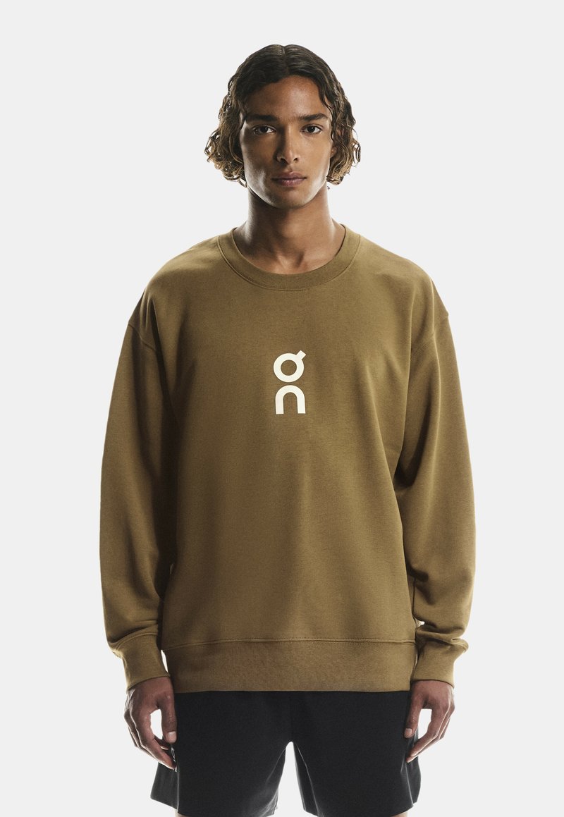Brun sweatshirt med lange ærmer, rund halsudskæring og et hvidt logo på brystet. Fremstillet af blødt, struktureret stof. Sammen med sorte shorts.