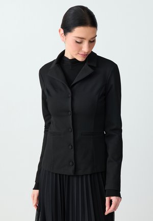 Kobieta z ciemnymi włosami upiętymi w niski kok, ubrana w dopasowany czarny zapinany na guziki żakiet, czarny golf i plisowaną spódnicę, patrząca w dół.