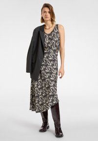 Abito a fantasia nero e crema con una silhouette fluida, abbinato a un blazer nero appoggiato sulla spalla e stivali bordeaux al ginocchio.