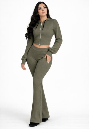 Femme aux longs cheveux foncés portant un sweat à capuche court zippé vert olive et un pantalon évasé, debout, main sur la hanche, sur fond blanc.