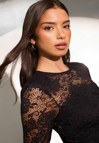 Melnā mežģīņu tunika ar ziedu rakstiem, caurspīdīgu tekstūru un garām piedurknēm. Modelei ir gari mati un riņķa auskari, ar vieglu dabīgu grimu.