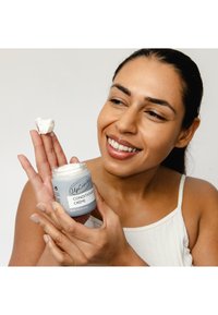 Przezroczysty słoik białego kremu odżywczego z etykietą "UpCircle". Krem jest trzymany na dłoni. Tło jest jednolite, a osoba ma ciemne, proste włosy.