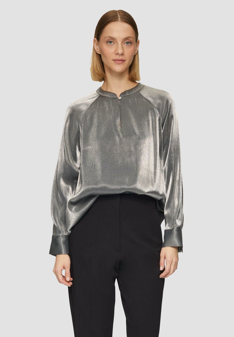 Zijdeachtige zilveren blouse met een ronde halslijn, knoopdetail en lange, losse mouwen. Gecombineerd met zwarte, high-waisted broeken.