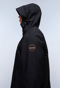 Sort hættetrøje lavet af letvægtsstof med et rundt logo-patch i orange nuancer på den øverste arm og sidelommer med lynlås.