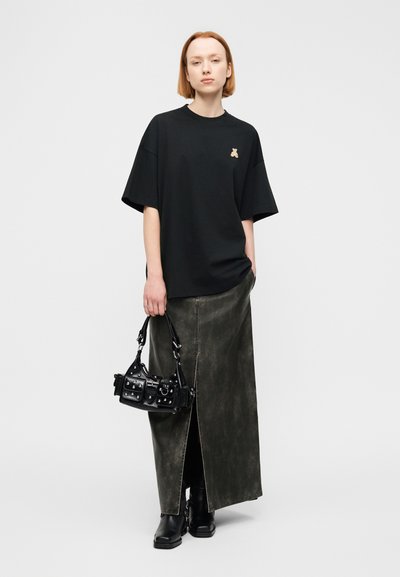 Μαύρο φαρδύ t-shirt με λογότυπο αρκούδας, συνδυασμένο με μια μακριά δερμάτινη φούστα σε σκουροπράσινο χρώμα με σκίσιμο μπροστά και μια μικρή μαύρη τσάντα χειρός.