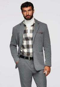 Grå blazer över en rutmönstrad skjorta med knappar, parat med matchande byxor. Anmärkningsvärda funktioner inkluderar en fickfyrkant och texturerad tyg.