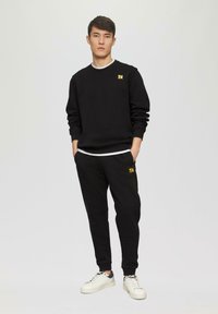 Schwarzes Sweatshirt mit langen Ärmeln und gerippten Bündchen, weiße Rundhalsausschnittverzierung, kombiniert mit passender schwarzer Jogginghose; beide tragen ein gelbes Logo.