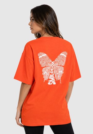 Kobieta zwrócona profilem, ubrana w pomarańczowy, oversize t-shirt z białym motylem wykonanym z wzorów odcisków palców i napisem „SMILODOX” na plecach.
