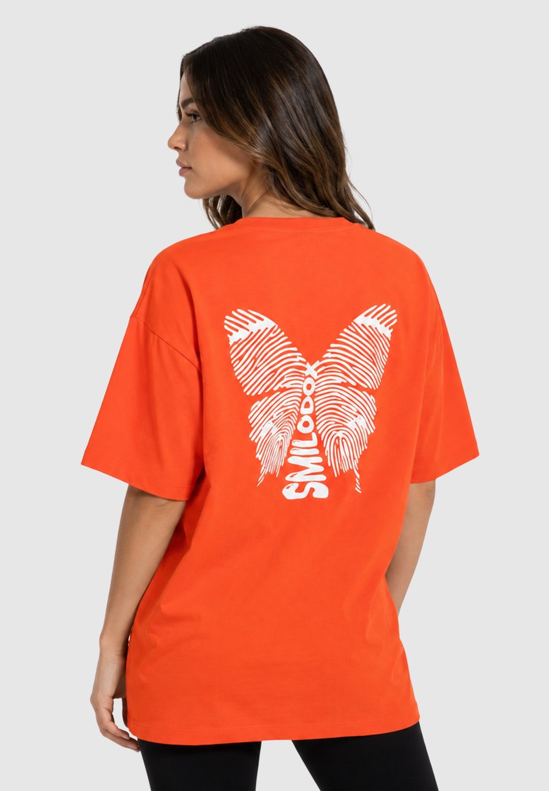 Kvinde set fra siden iført en orange oversized t-shirt med en hvid sommerfugl lavet af fingeraftryksmønstre og ordet "SMILODOX" på ryggen.