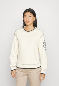 Pullover in crema chiaro con tessuto testurizzato, bordo nero lungo il colletto e i polsini, e un patch con logo sulla manica sinistra.