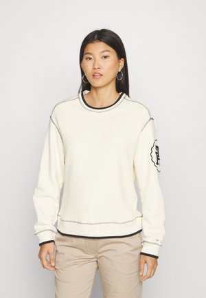 Pullover in crema chiaro con tessuto testurizzato, bordo nero lungo il colletto e i polsini, e un patch con logo sulla manica sinistra.