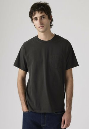Jonge man met kort donker haar, gekleed in een effen zwart T-shirt met korte mouwen en donkerblauwe spijkerbroek, staand tegen een effen achtergrond.