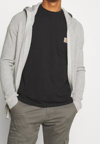 Šedý kardigan s vroubkovanou texturou, nošený přes černé tričko s kapsou na prsou, v kombinaci s olivovými cargo kalhotami. Jednoduchý a funkční design.