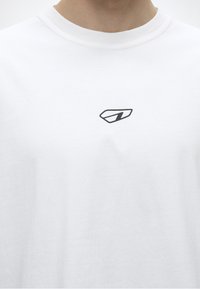 Κοντινό πλάνο ενός λευκού T-shirt με στρογγυλή λαιμόκοψη και ένα μικρό μαύρο αφηρημένο γεωμετρικό λογότυπο στην αριστερή πλευρά του στήθους.