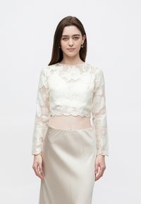 Nuori nainen seisoo valkoisessa kukallisessa pitsisessä pitkähihanaisessa crop-topissa ja korkeavyötäröisessä beigenvärisessä satiinihameessa yksiväristä taustaa vasten.