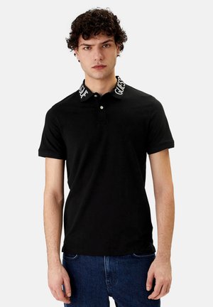 Giovane uomo con capelli ricci che indossa una polo nera con la scritta "GUESS" sul colletto e jeans blu, in piedi davanti a uno sfondo neutro.