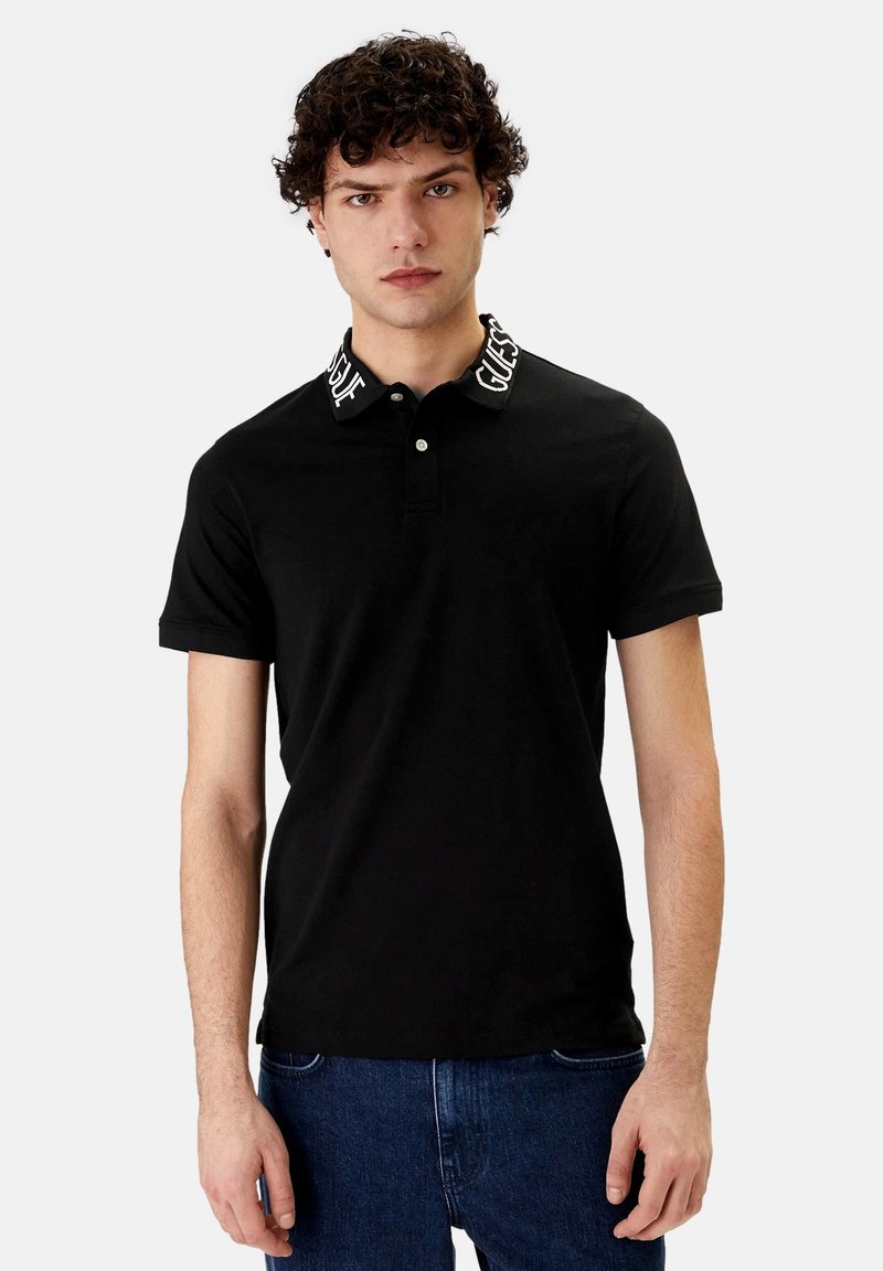 Giovane uomo con capelli ricci che indossa una polo nera con la scritta "GUESS" sul colletto e jeans blu, in piedi davanti a uno sfondo neutro.