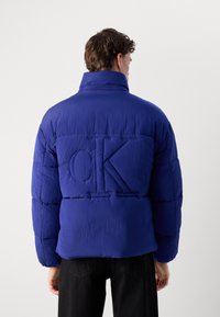 Blå pufferjakke med høy krage, teksturert stoff, quiltet design og en preget "CK"-logo på ryggen. Sorte bukser synlige.