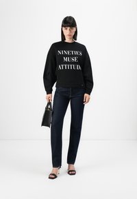 Felpa nera con testo bianco "NINETIES MUSE ATTITUDE", abbinata a jeans in denim scuro, borsa nera e sandali neri con tacco alto.