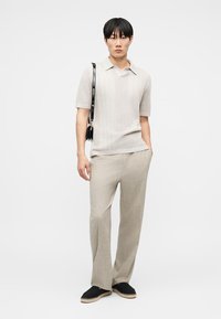 Νεαρός άνδρας με μπεζ πλεκτό polo με κοντά μανίκια και παντελόνι, μαύρη τσάντα ώμου και μαύρα εσπαντρίγιες, στέκεται μπροστά σε λευκό φόντο.