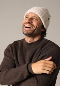 Bruine geribbelde trui met een hoge nek, gecombineerd met een lichtbeige gebreide muts. De muts heeft een klein merklabel.