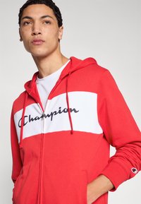 Κόκκινο φούτερ με φερμουάρ, με λευκή επιφάνεια στο στήθος που φέρει μαύρο λογότυπο "Champion". Κατασκευασμένο από απαλό υλικό, με κορδόνια και πλαϊνές τσέπες.