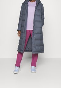 Manteau doudoune gris foncé avec capuche, doté de boutons-pression, associé à une chemise à manches longues violet clair et un pantalon rose. Des chaussures bleu clair viennent compléter l'ensemble.